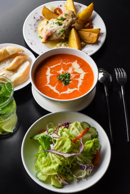 Mittagsset aus Tomatencremesuppe, Salat, Hähnchen mit Kartoffeln, Limonade auf schwarzem Hintergrund