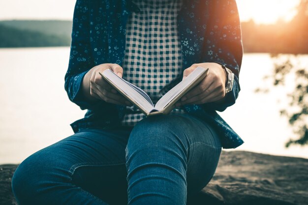 Foto mittelabschnitt einer frau mit einem buch, während sie bei sonnenuntergang gegen den see sitzt