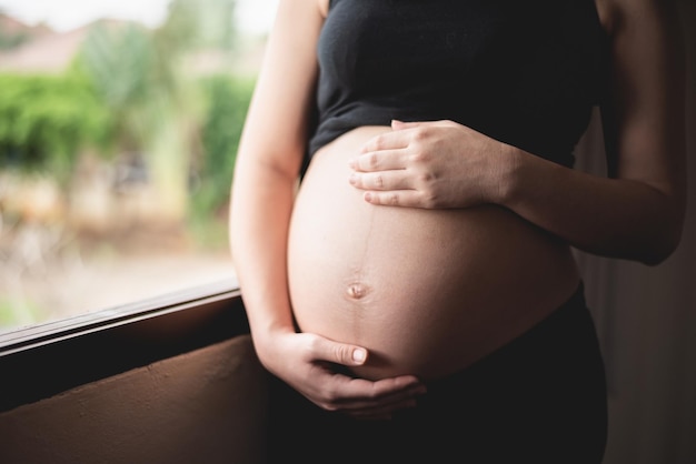 Mittelabschnitt einer schwangeren Frau, die den Bauch am Fenster berührt