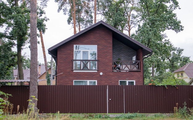 Mittelkleines braunes Privathaus mit Wellpappezaun im grünen Wald im Sommer. Landhaus im Hintergrund des Waldes. Vermietung von Ferienhäusern außerhalb der Stadt für die Ferien.