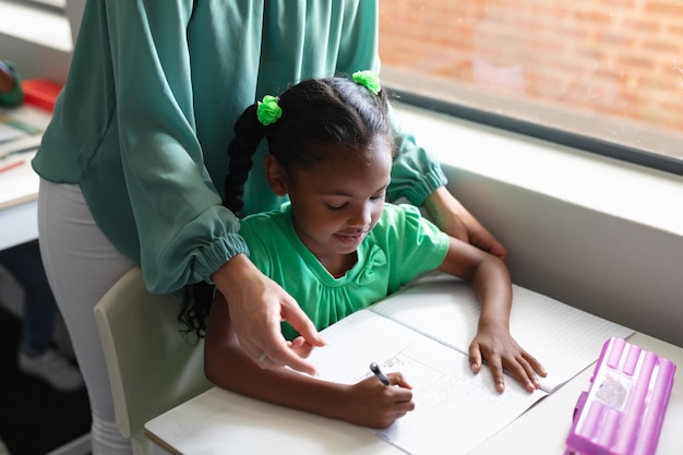 Mittelschnitt einer jungen weißen Lehrerin, die einem afroamerikanischen Schulmädchen hilft, das am Schreibtisch studiert.