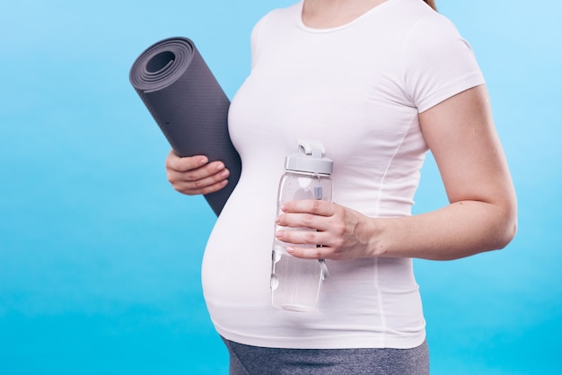 Mittelteil der aktiven schwangeren frau, die flasche wasser und fitness-teppich hält, während über blauer wand steht