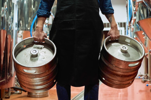 Mittelteil des männlichen Arbeiters der Bierproduktionsanlage im Vorfeld, der zwei Zisternen trägt, während er sich vor der Kamera entlang Reihen riesiger Stahltanks bewegt