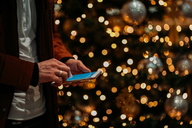 Mittelteil des Mannes, der intelligentes Telefon gegen belichteten Weihnachtsbaum verwendet.