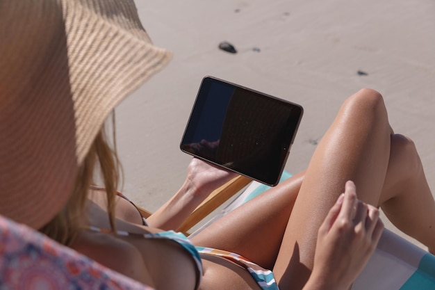 Mittelteil einer kaukasischen Frau, die ein digitales Tablet nutzt, während sie auf einem Liegestuhl am Strand sitzt. Sommer-Strandurlaubskonzept.