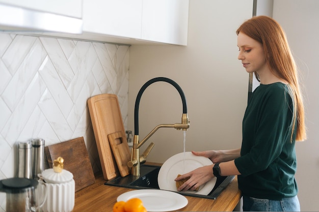 Mittlere Aufnahme einer attraktiven rothaarigen jungen Frau, die Geschirr in der Spüle mit Schwamm in der Küche wäscht