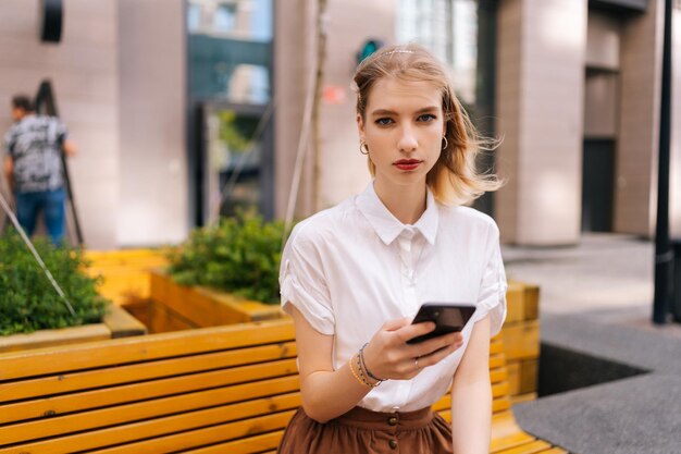 Mittlere Aufnahme einer ernsthaften, hübschen jungen Frau, die auf einer gelben Bank auf der Straße der Stadt sitzt und in die Kamera schaut, die ein Handy in der Hand auf dem Hintergrund des Gebäudes hält