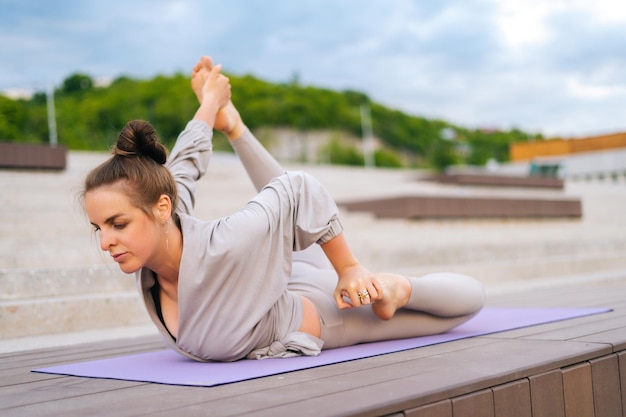 Mittlere Aufnahme einer flexiblen jungen Yogini-Frau, die an Sommertagen im Freien im Stadtpark Yoga-Übungen auf einer Fitnessmatte praktiziert Ruhige Frau mit schönem Körper in Sportbekleidung, die draußen Asana-Pose aufführt