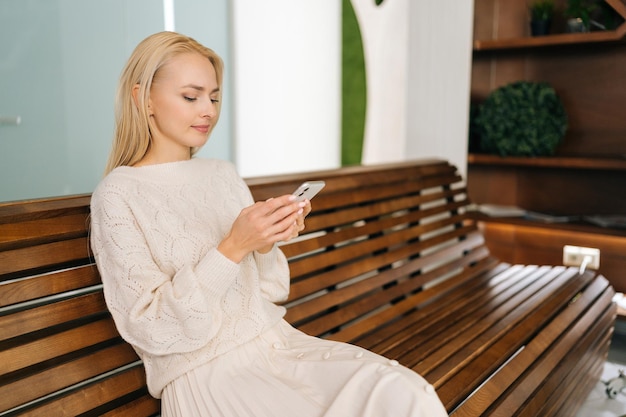 Mittlere Aufnahme einer jungen blonden Frau, die mit ihrem Smartphone auf einer Holzbank sitzt und auf den Bildschirm ihres Mobiltelefons blickt und sich an der Nutzung mobiler Apps erfreut