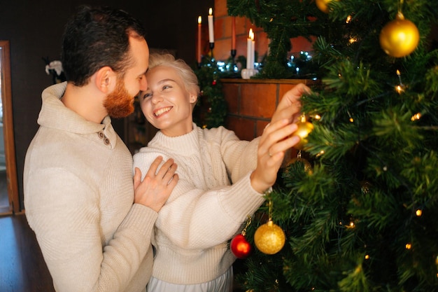 Mittlere Aufnahme eines charmanten jungen verliebten Paares, das den Weihnachtsbaum mit glänzenden Kugeln in der Nähe des Kamins in einem gemütlichen dunklen Wohnzimmer schmückt