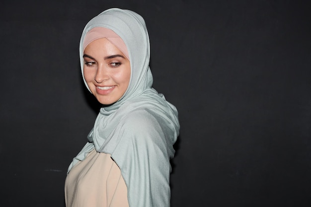 Mittlere Nahaufnahme Studioporträt eines charmanten muslimischen Mädchens mit hellblauem Hijab, das gegen bla steht