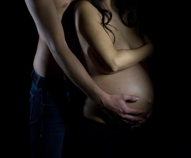 Foto mittlerer abschnitt eines hemdlosen mit händen auf dem bauch einer schwangeren frau auf schwarzem hintergrund