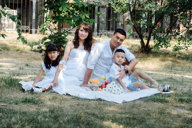 Mittleres Distanzporträt einer asiatischen Familie in weißer Kleidung, die in einem Park picknickt und die letzten warmen Tage des frühen Herbstes genießt. Eltern und ihre Kinder in einem Baumschatten.