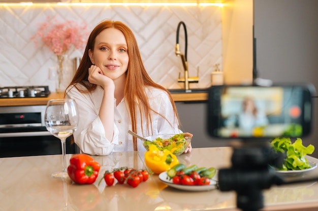 Mittleres Porträt einer lächelnden, attraktiven rothaarigen Vloggerin, die einen Video-Food-Blog über das Kochen vor der Kamera eines Mobiltelefons dreht, das in der Küche sitzt, mit modernem, hellem, selektivem Fokus