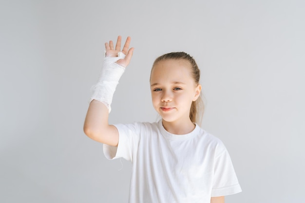 Mittleres Porträt eines fröhlichen kleinen Mädchens mit gebrochener Hand, eingewickelt in einen weißen Gipsverband, grüßt den hebenden Arm und blickt in die Kamera
