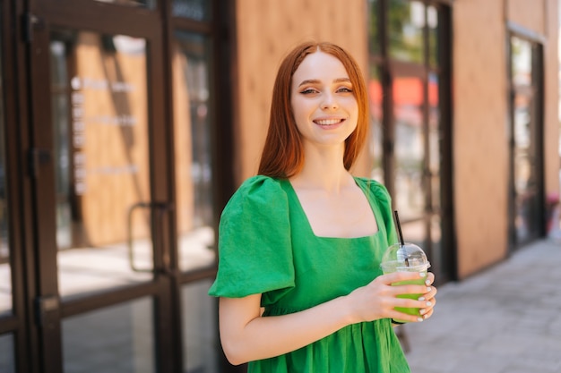 Mittleres Schussporträt einer fröhlichen, attraktiven jungen Frau, die an einem sonnigen Sommertag einen coolen Cocktail auf der Stadtstraße hält und die Kamera anschaut. Hübsche Dame, die kaltes Getränk durch Stroh trinkt.