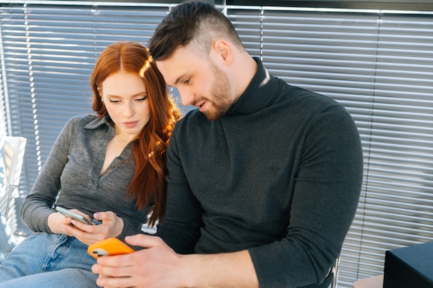 Mittleres Schussporträt eines gutaussehenden jungen Mannes, der sein Handy einer attraktiven Frau zeigt, die in der Warteschlangenreihe in der Bürolobby sitzt. Glückliches junges Paar, das auf der Treppe im Dachboden sitzt und Handy teilt