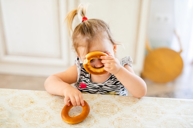 Miúdo pequeno da criança que tem o almoço na cozinha ensolarada morna. Menina loira com rabo de cavalo engraçado brincar com dois bagels saborosos