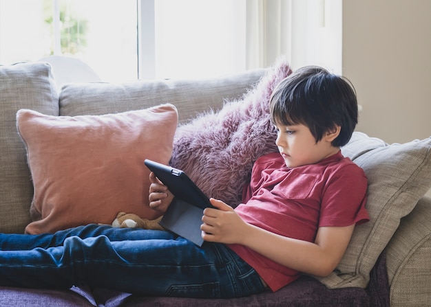 Mixed Race Kid sitzt auf dem Sofa und schaut sich Cartoons auf dem Tablet an oder spielt Spiele auf dem Touchpad.