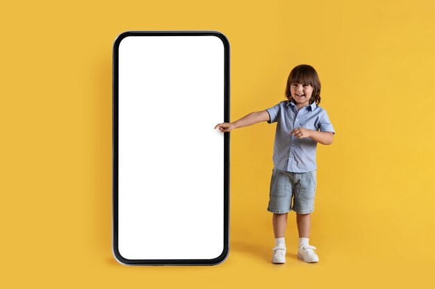 Mobile App für pädagogische Kinder Fröhlicher, positiver kleiner Junge, der auf ein großes Smartphone mit schwarz-weißem Bildschirm zeigt