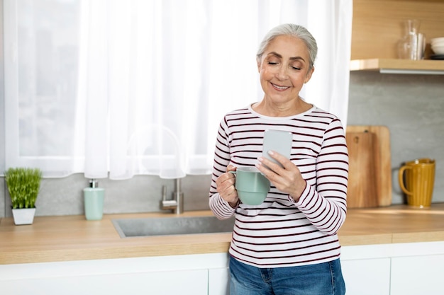 Mobilkommunikation Lächelnde ältere Dame, die sich in der Küche mit Smartphone und Kaffee entspannt