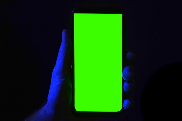 Mobiltelefon in der Hand eines jungen Mannes Verwenden Sie ein Smartphone mit grünem Bildschirm auf rotem, blauem Neon, das dunklen Hintergrund beleuchtet