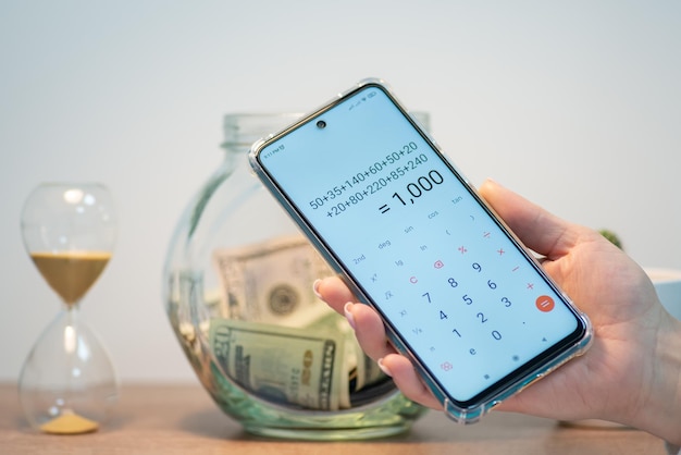 Mobiltelefon mit Berechnung auf dem Bildschirm, Glasgefäß mit Dollar-Banknoten und Sanduhr auf dem Tisch im Hintergrund. Geldberechnungs- oder Sparplanungskonzepte für Finanzinvestitionen