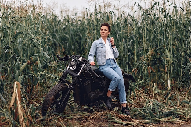Moça bonita atraente vestindo roupa urbana casual com motocicleta de estilo clássico. Motociclista feminina ao ar livre. No fundo da natureza ou da grama. Conceito de moto