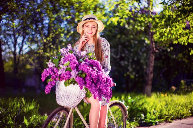Moça bonita com bicicleta do vintage e flores na cidade na luz solar ao ar livre.