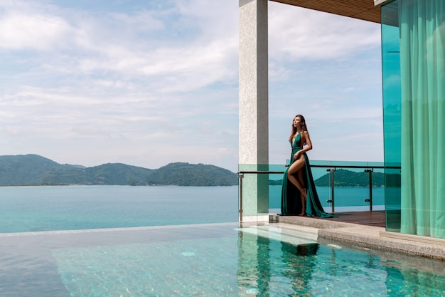 Moça bonita em um vestido verde longo posando perto de uma piscina ao ar livre com vista para o mar e montanhas verdes