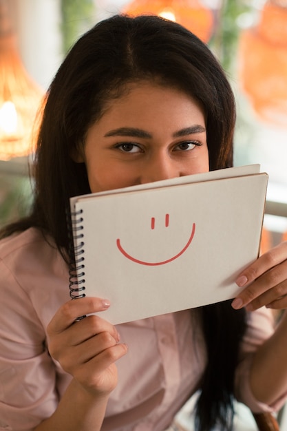 Foto moça bonita olhando para a câmera enquanto tem o rosto escondido atrás do sorriso desenhado na página do caderno branco