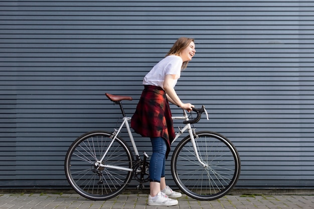 Foto moça bonita que está com uma bicicleta branca no fundo da parede listrada cinza