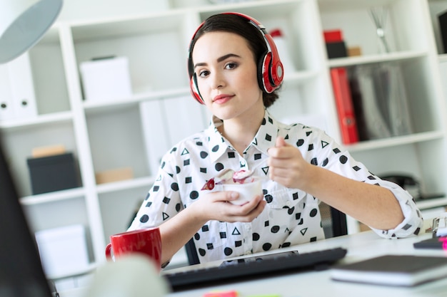 Moça bonita que senta-se nos fones de ouvido na mesa no escritório que come o iogurte com enchimento vermelho.