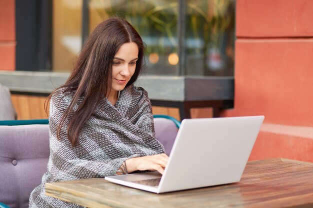 Moça bonita senta-se na frente do laptop aberto, escreve e-mail, usa internet de alta velocidade, embrulhada em xadrez, posa na cafeteria ao ar livre