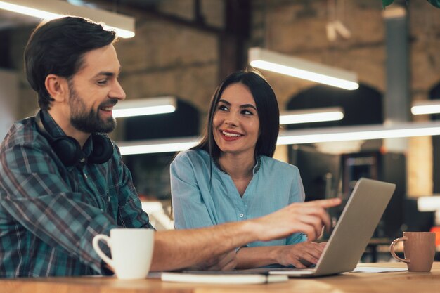 Moça bonita sorrindo e olhando para o jovem ao seu lado apontando para uma tela de laptop no escritório