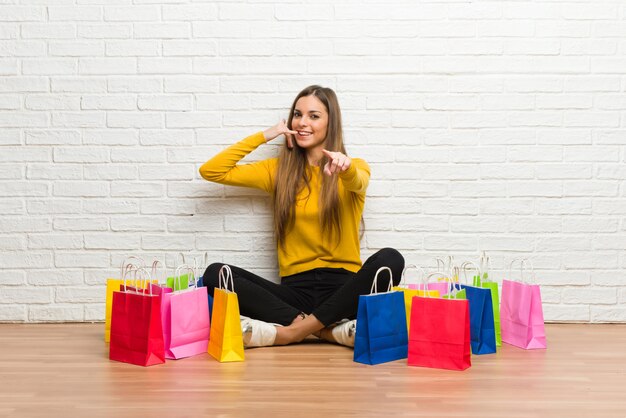 Moça com muitos sacos de compras, fazendo o gesto do telefone e apontando a frente