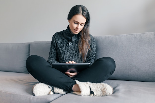 Moça na camisola de malha em casa no sofá com tablet digital