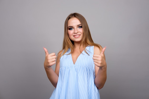 Moça no vestido azul que mostra os polegares, isolado no cinza