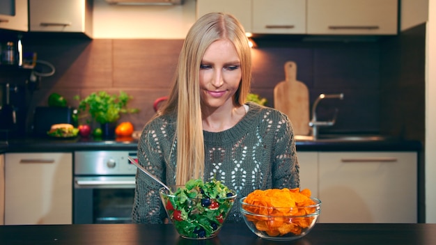 Moça preferindo salada crocante. Mulher jovem e bonita escolhendo comer batata frita saudável para o jantar enquanto está sentado à mesa na cozinha elegante e ela come essa salada.