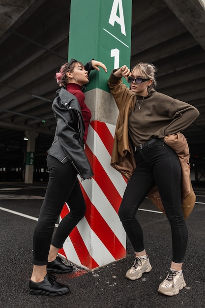 Moças elegantes e lindas com roupas jeans vintage da moda e jaquetas de couro, posando em um estacionamento na cidade