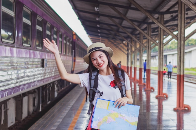 Mochila asiática mujer viajero que tiene un mapa local genérico y agita la mano en la estación de tren