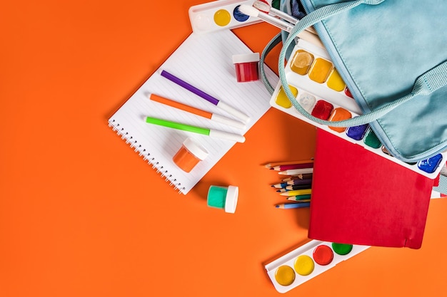 Una mochila azul con pinturas de papelería de diferentes colores sobre un fondo naranja