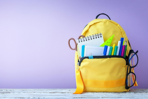 Mochila escolar amarela completa com óculos de papelaria e canetas de notas na mesa em fundo roxo Conceito de volta à escola