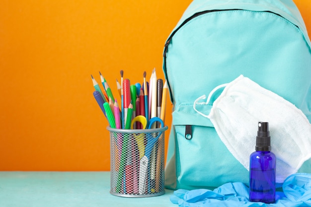 Mochila escolar azul con máscara, desinfectante para manos, guantes y papelería en la mesa. Nueva vida normal.