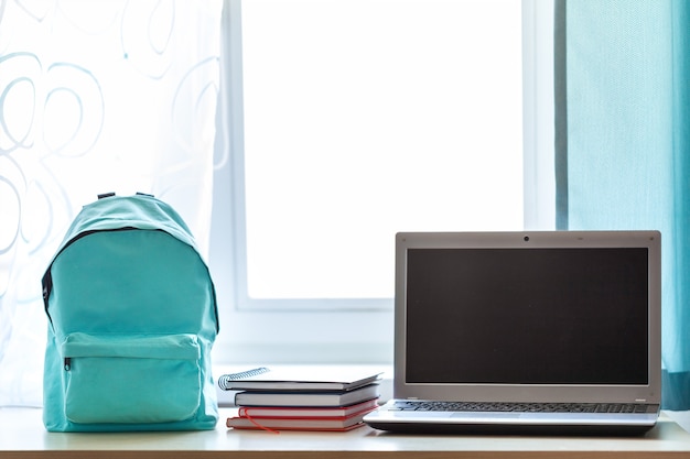 Mochila escolar azul con útiles escolares y computadora en la mesa en la habitación de los niños frente a la ventana en un día soleado