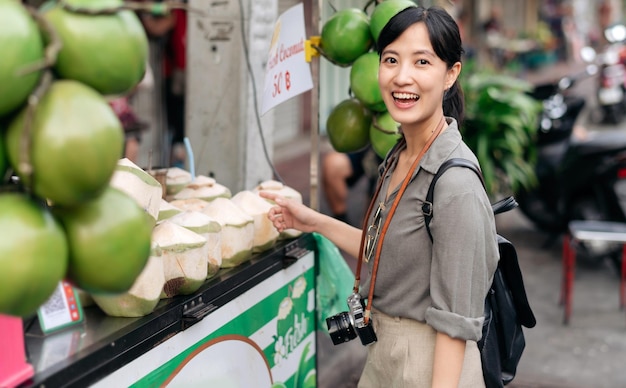 Mochila jovem asiática feliz que viaja em frente a uma loja de suco de coco no mercado de comida de rua da cidade chinesa em Bangkok Tailândia Viajante verificando ruas laterais