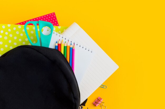 Foto mochila preta e material escolar sobre um fundo amarelo com espaço de cópia, configuração plana.