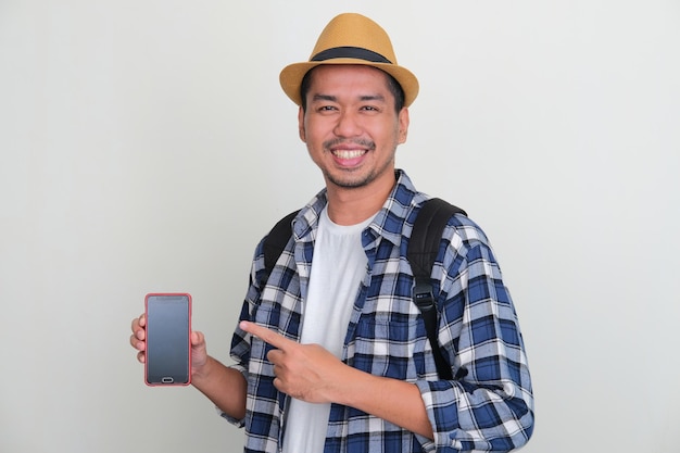 Mochileiro asiático sorrindo para a câmera enquanto aponta para a tela do telefone em branco que ele segura