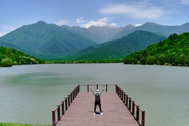 Mochileiro de homem viajante sozinho no cais e olhando para o lago e as montanhas. desfrutando de um belo momento de liberdade e de uma atmosfera serena e tranquila na natureza. vista traseira