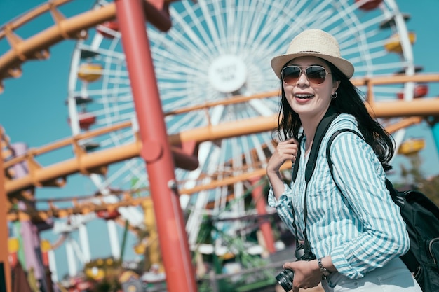Mochileiro de jovem chinesa asiática feliz se divertindo no feriado de férias de primavera. Mulher alegre na excursão de um dia de férias na roda gigante. mochileiro feliz em óculos de sol e chapéu de palha carrega câmera sorrindo